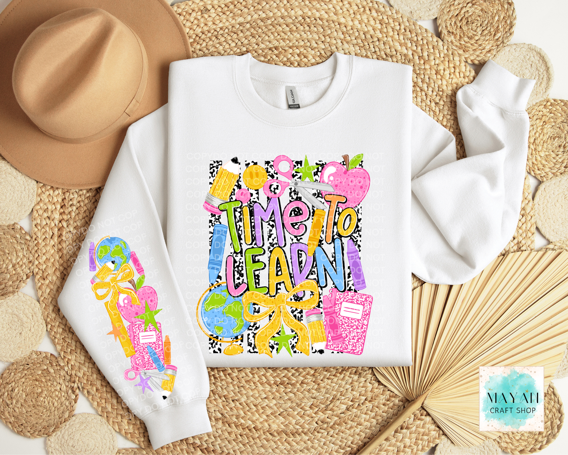 A white crewneck sweatshirt with colorful school-themed graphics, including the text 'Time To LEARN' and illustrations of books, pencils, a globe, and a notebook. The sweatshirt is displayed in a flat-lay arrangement on a woven mat with a tan hat, a folded white towel, and a dried palm leaf.