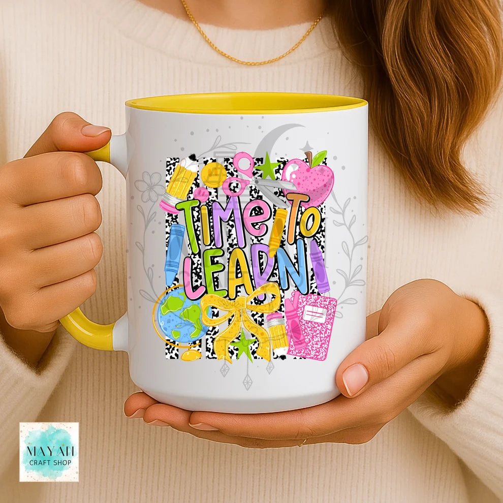 A person holds a white ceramic mug with a yellow interior and handle. The mug features a colorful 'Time to Learn' school-themed graphic with apples, books, and a globe.