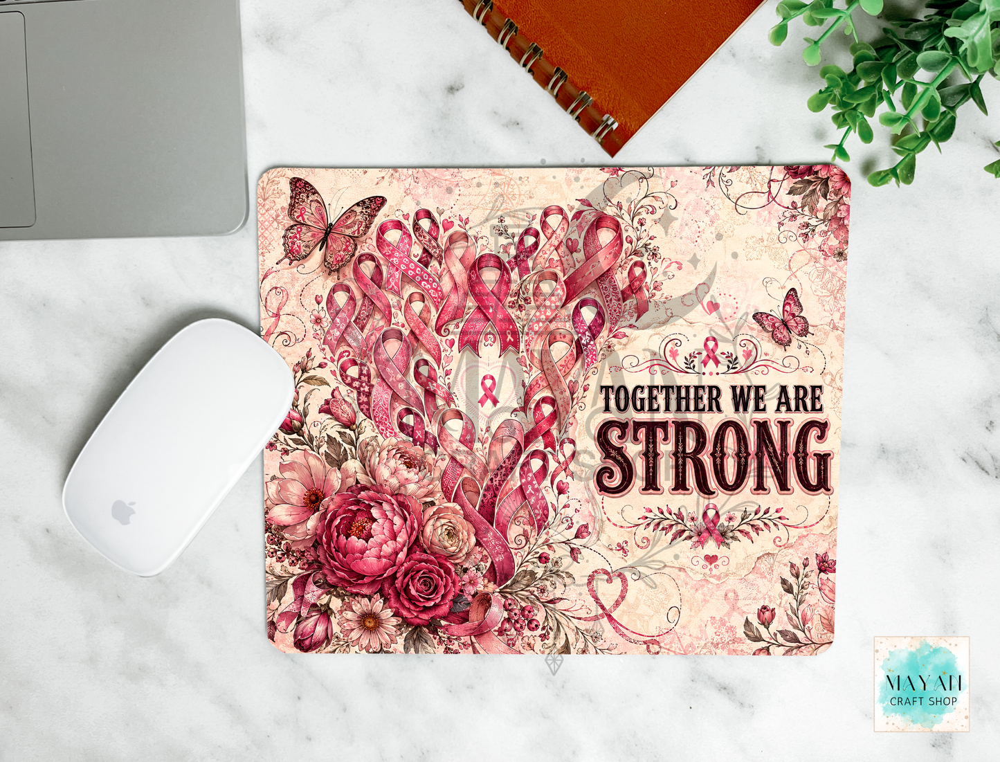 A mouse pad with a pink and red floral design, butterflies, and the text 'TOGETHER WE ARE STRONG' is shown on a white marble surface with a laptop mouse, notebook, and plant nearby.