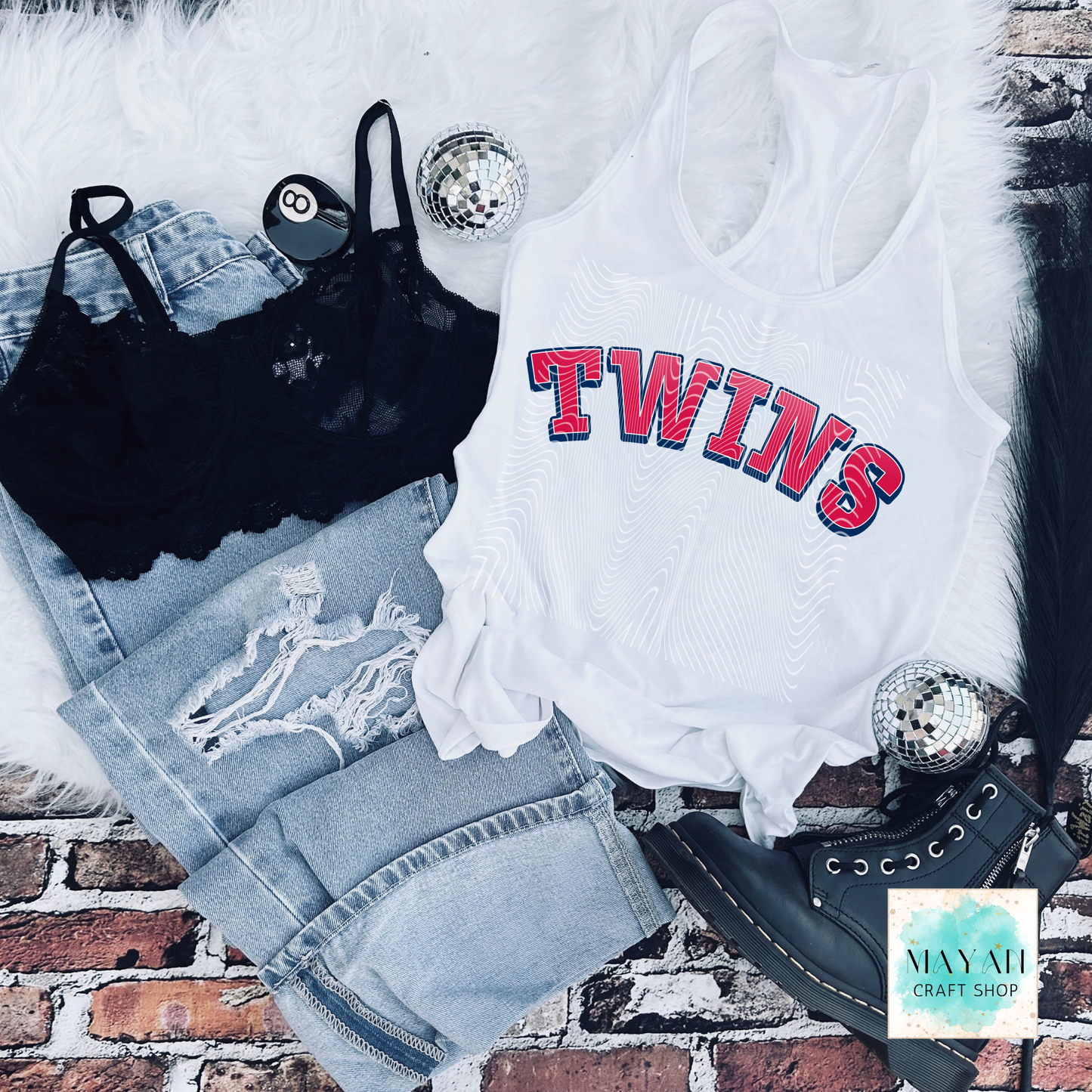 White tank top with a baseball themed text, black bralette, denim shorts, and black shoes on a brick surface.