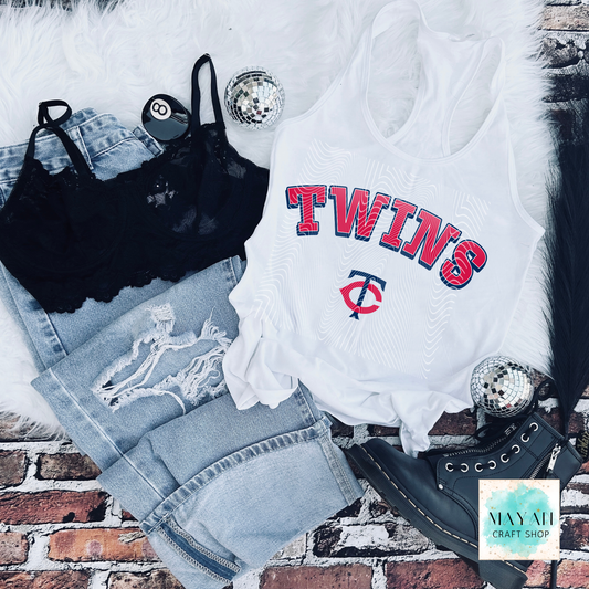 White tank top with a baseball themed print, black bralette, denim shorts, and black shoes on a brick surface.