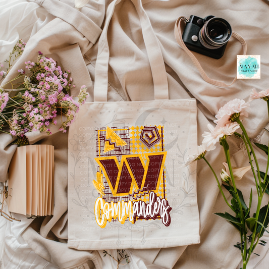 A cream canvas tote bag with a graphic design featuring a large letter and a football team logo in yellow and maroon, styled in a flat lay with flowers, a camera, and a notebook on a beige fabric background.