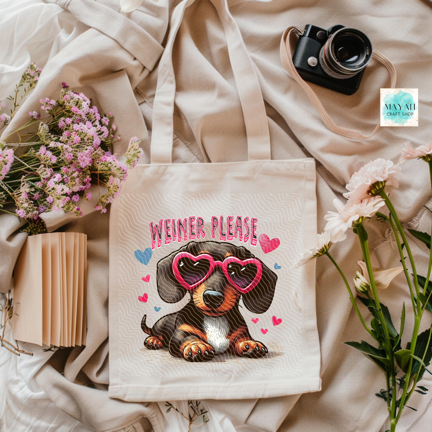 Tote bag with a dog graphic and ' Weiner Please' text on a soft fabric background with flowers and a camera.