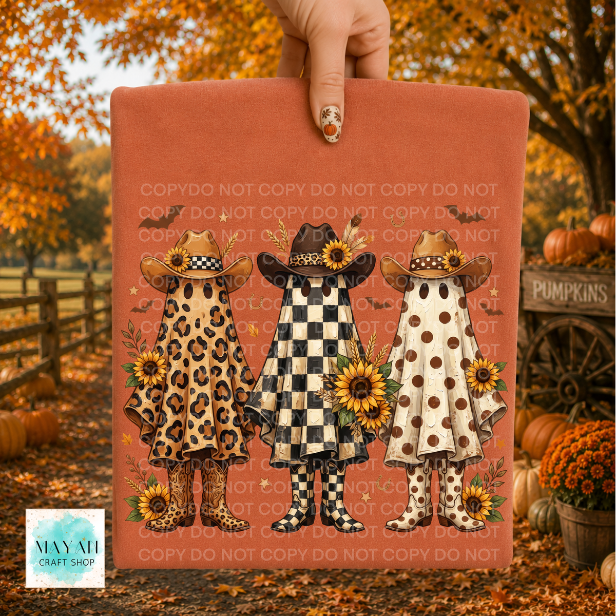 A shirt held by a hand, featuring three illustrated girls in autumn outfits with sunflowers and rain boots. The sign is photographed outdoors in a fall-themed setting with pumpkins, orange leaves, and a fence in the background.