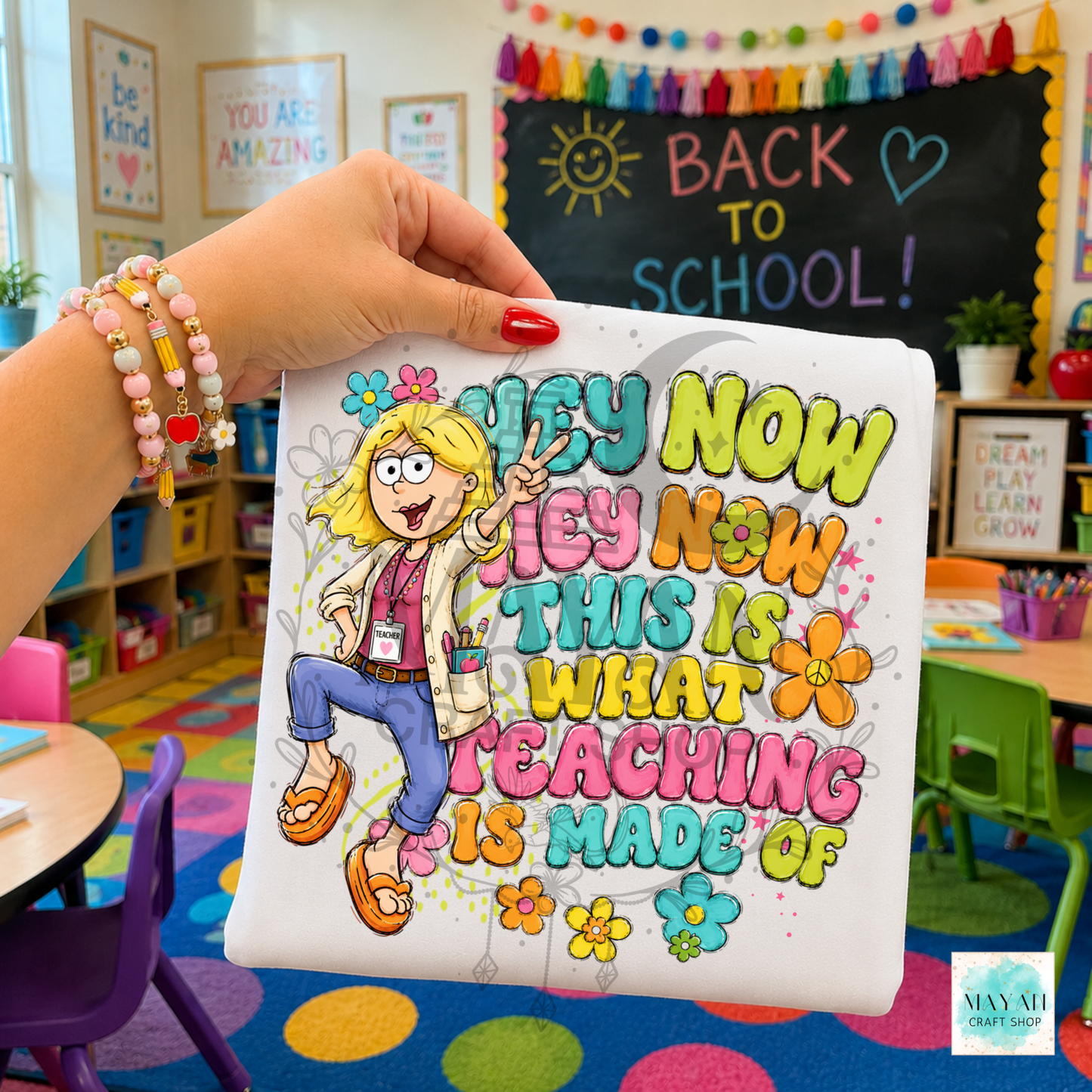 A hand holds a white T-shirt featuring a colorful cartoon graphic of a smiling blonde woman making a peace sign, surrounded by flowers and the text 'what teaching is made of.' The background shows a decorated classroom with a chalkboard that says 'BACK TO SCHOOL!' and colorful furniture.