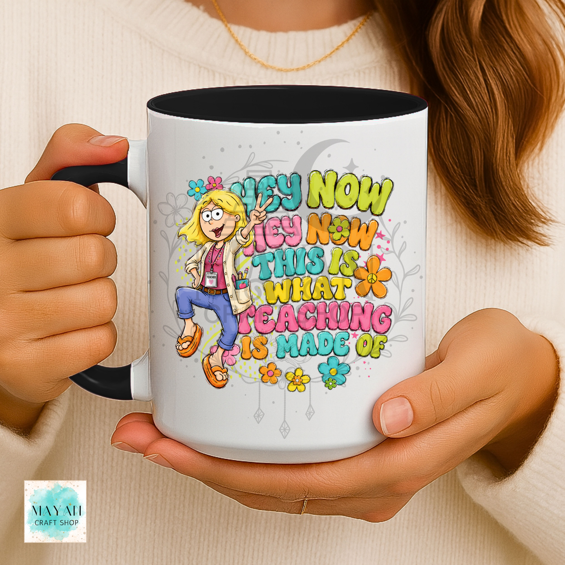 A person holds a white ceramic mug with a black handle and interior. The mug features a colorful cartoon illustration of a blonde woman and the text 'HEY NOW THEY NOW THIS IS WHAT TEACHING IS MADE OF' in playful multicolored lettering.