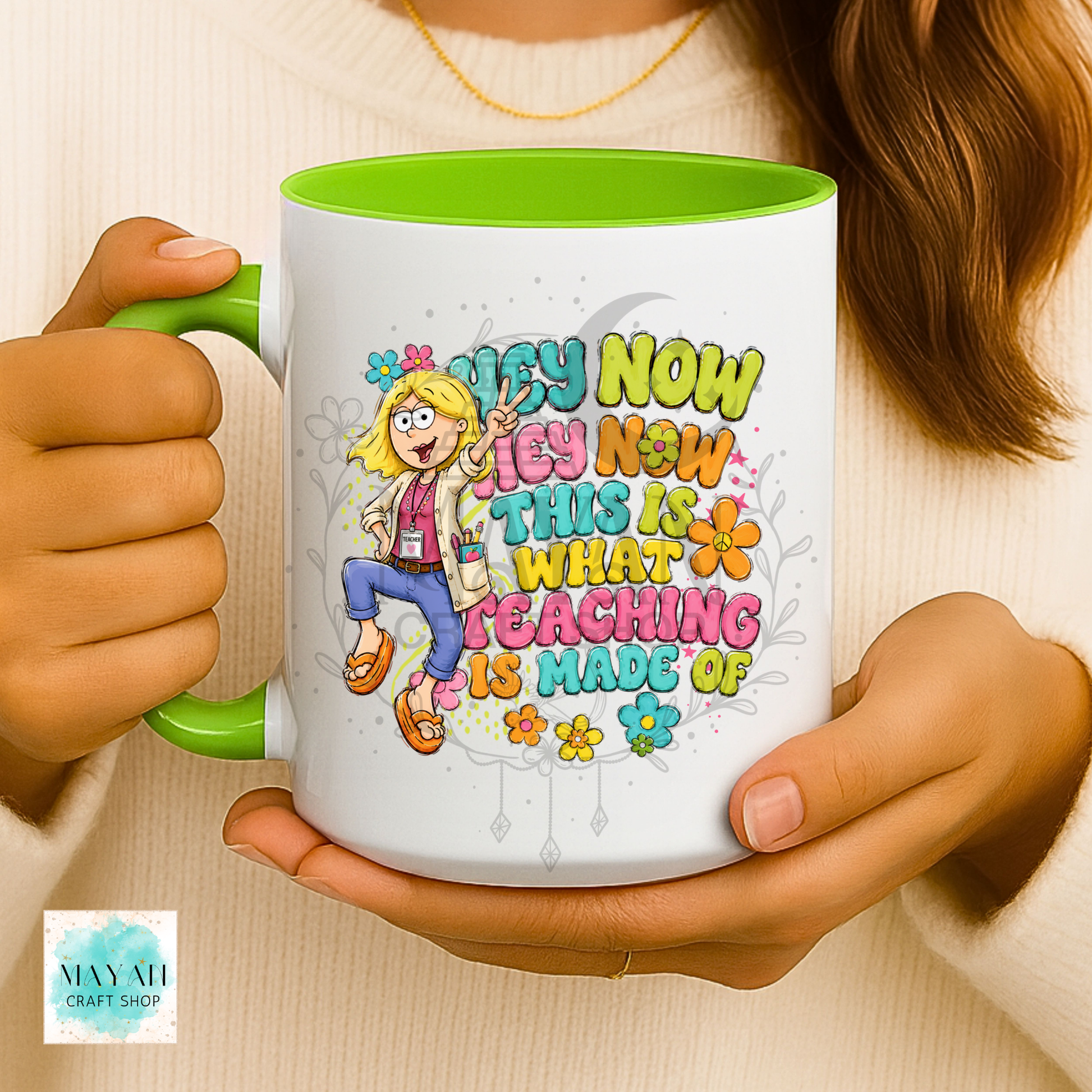 A person holds a white ceramic mug with a bright green handle and interior. The mug features a colorful cartoon illustration of a smiling teacher and the text 'TEACH NOW TEACH NOW THIS IS WHAT TEACHING IS MADE OF' in playful multicolored lettering.