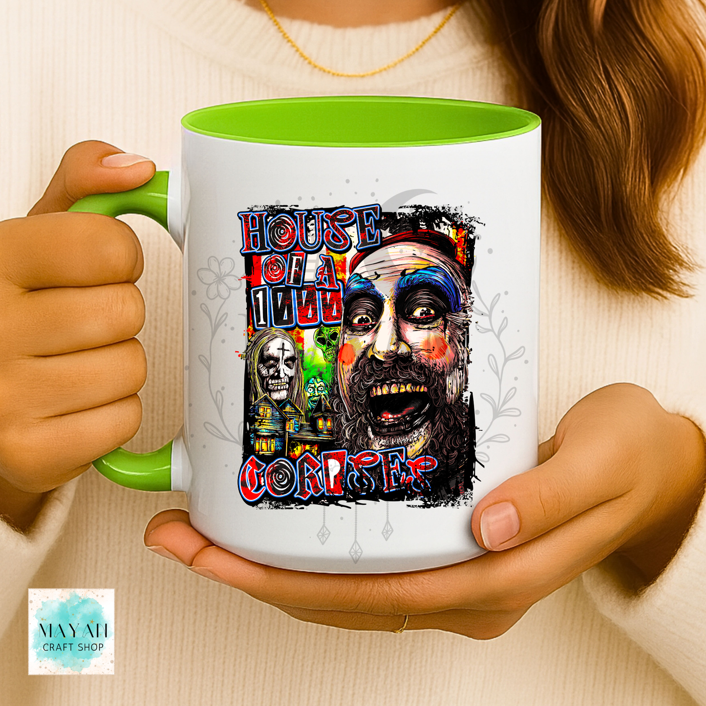 A person holds a white ceramic mug with a bright green handle and interior. The mug features a colorful horror-themed graphic with a text and a stylized illustrated face.