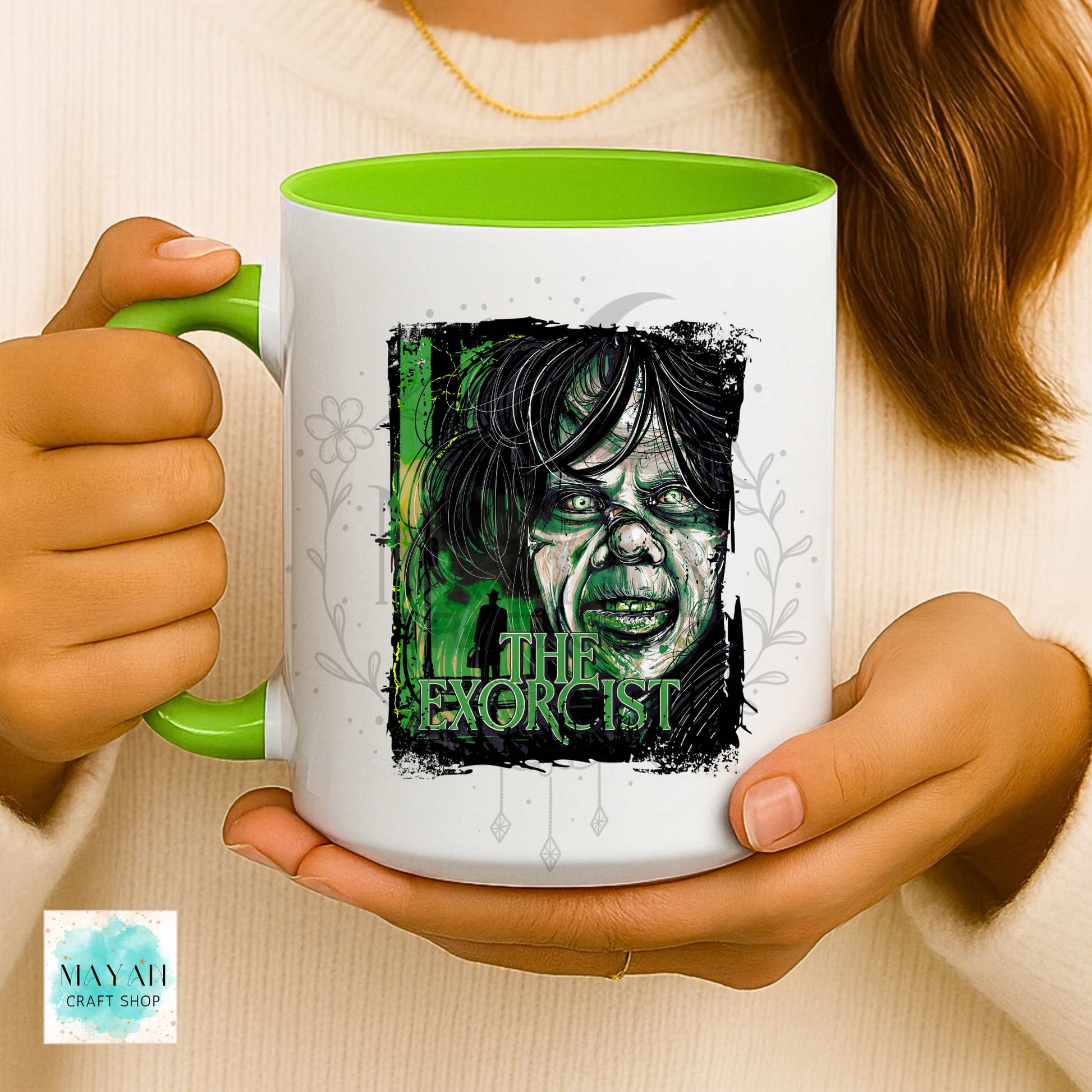 A person holds a white ceramic mug with a bright green handle and interior. The mug features a stylized green-and-black illustrated face with a text and faint floral line art.