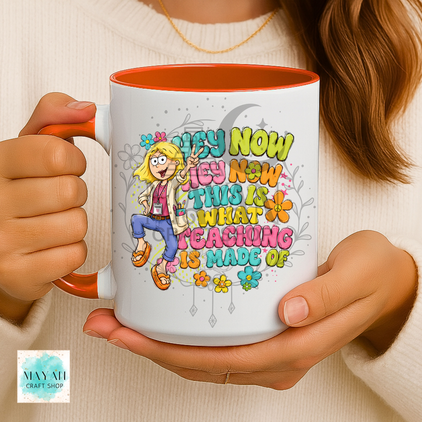 A person holds a white ceramic mug with orange accents and a colorful cartoon graphic that reads 'HEY NOW THIS IS WHAT TEACHING IS MADE OF.' The mug is shown against a cream sweater background.