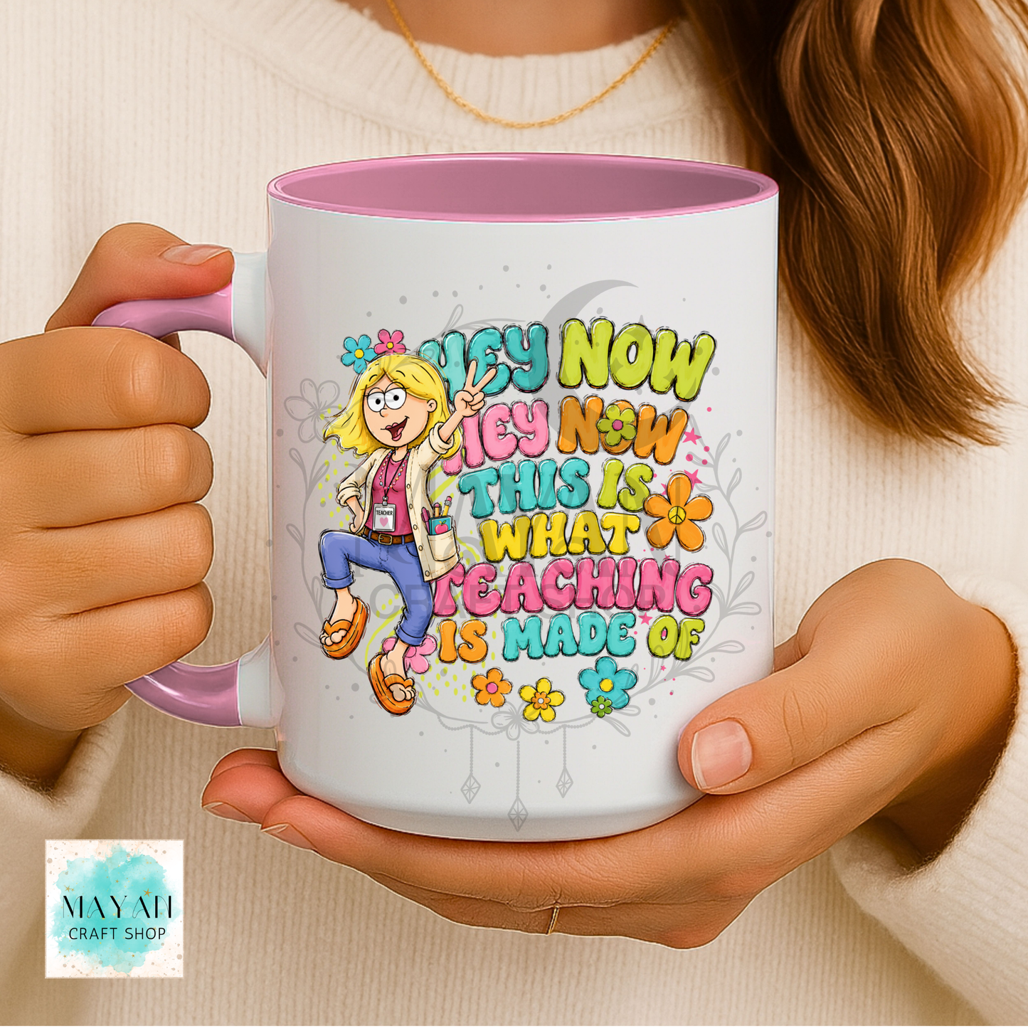 A person holds a white ceramic mug with a pink interior and handle. The mug features a colorful cartoon illustration of a blonde woman and the text 'HEY NOW THIS IS WHAT TEACHING IS MADE OF' in playful multicolor lettering.