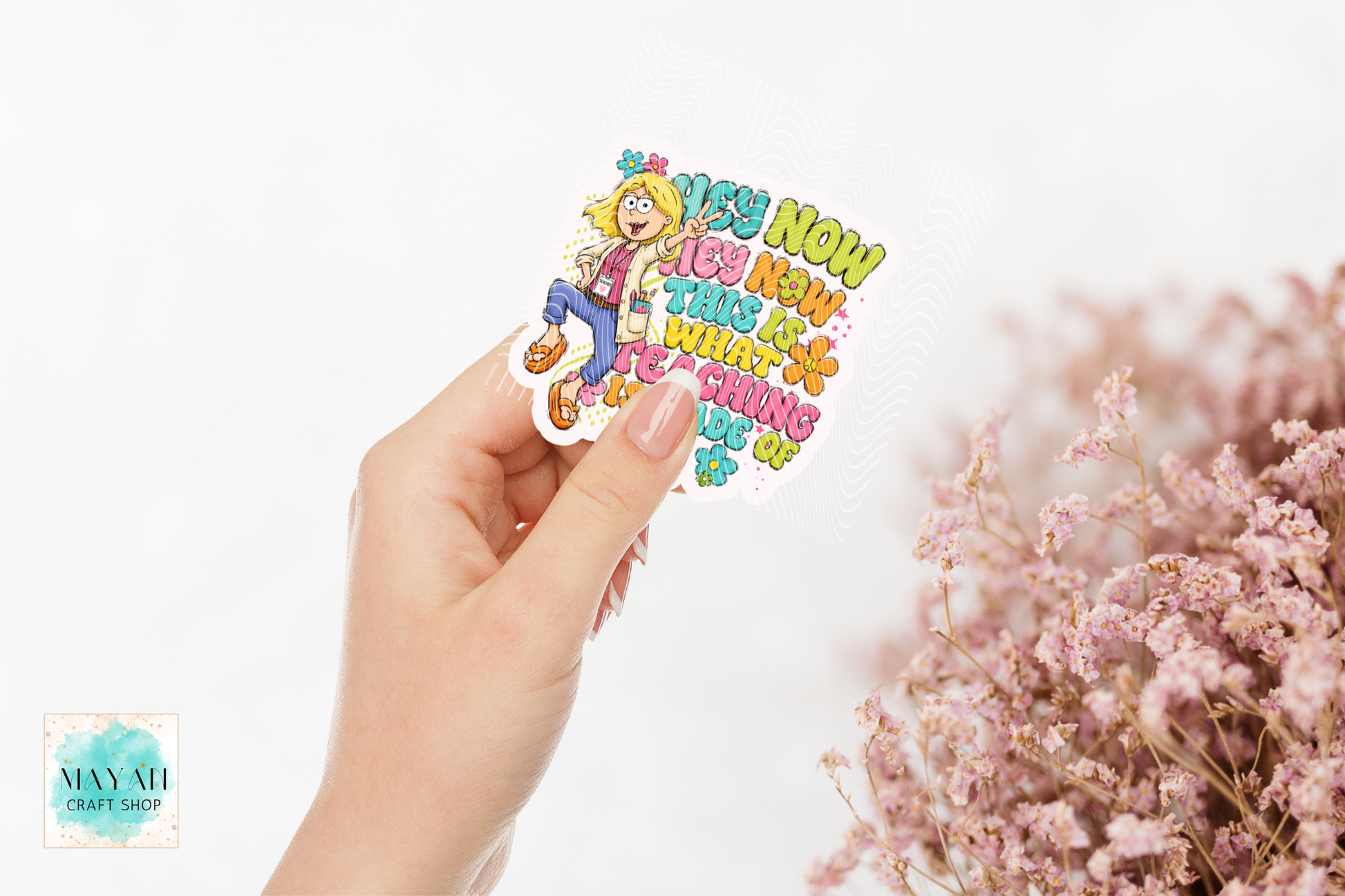 A hand holds a die-cut sticker featuring a cartoon girl and colorful retro text that reads 'HEY NOW I'M KNOCKED WEARING MY NEW SKINNY Jumpsuit' against a white background with blurred pink flowers on the right.