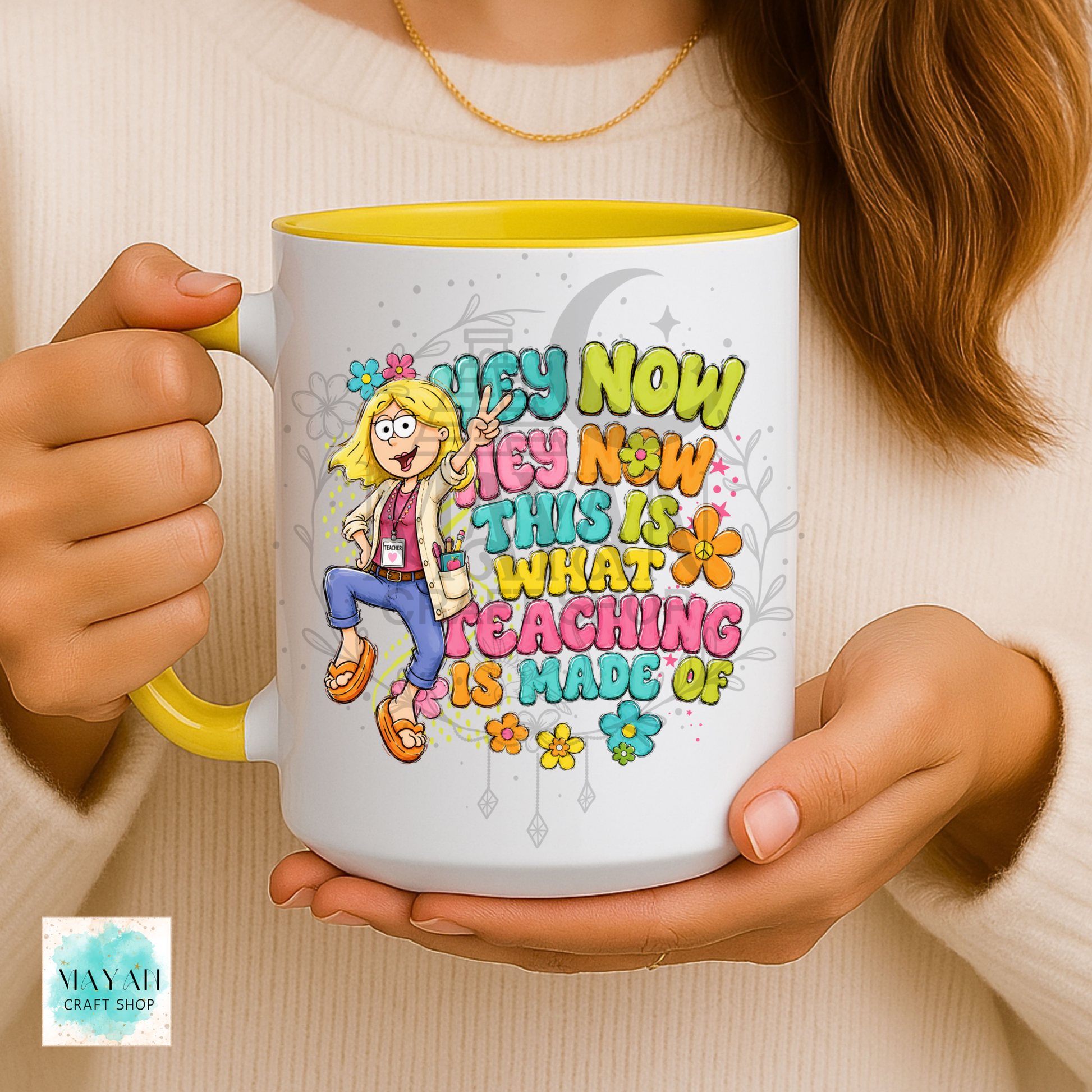 A person holds a white ceramic mug with a yellow interior and handle. The mug features a colorful cartoon illustration of a female teacher and the text 'TEACH NOW THIS IS WHAT TEACHING IS MADE OF' in playful multicolored lettering.