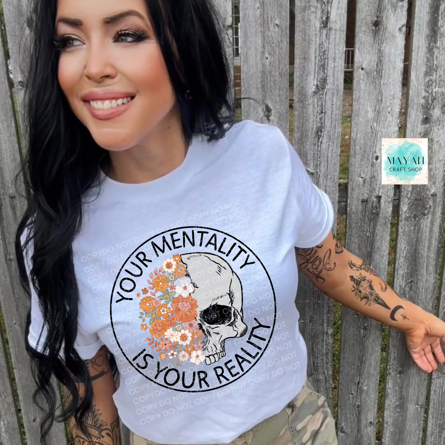 Woman wearing a white t-shirt with a skull and floral design, text 'Your Mentality is Your Reality', standing against a wooden fence.