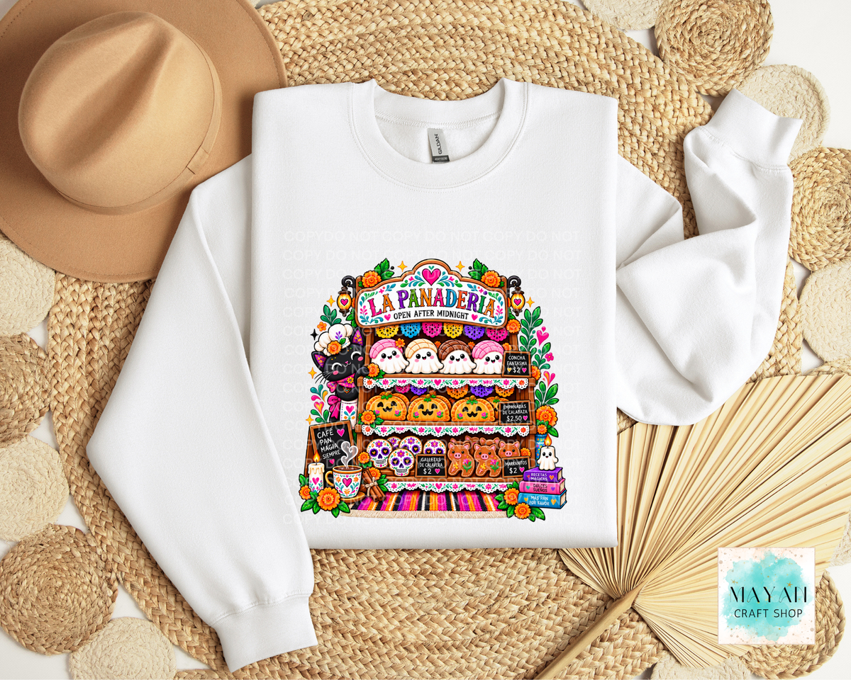 A white crewneck sweatshirt laid flat on a woven straw mat, featuring a colorful graphic print of a Halloween-themed market stall labeled 'La Panaderia' with cute characters and pumpkins. A tan hat, woven mat.