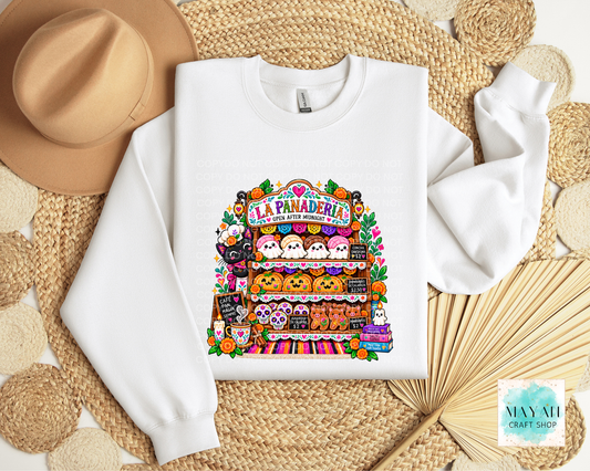 A white crewneck sweatshirt laid flat on a woven straw mat, featuring a colorful graphic print of a Halloween-themed market stall labeled 'La Panaderia' with cute characters and pumpkins. A tan hat, woven mat.