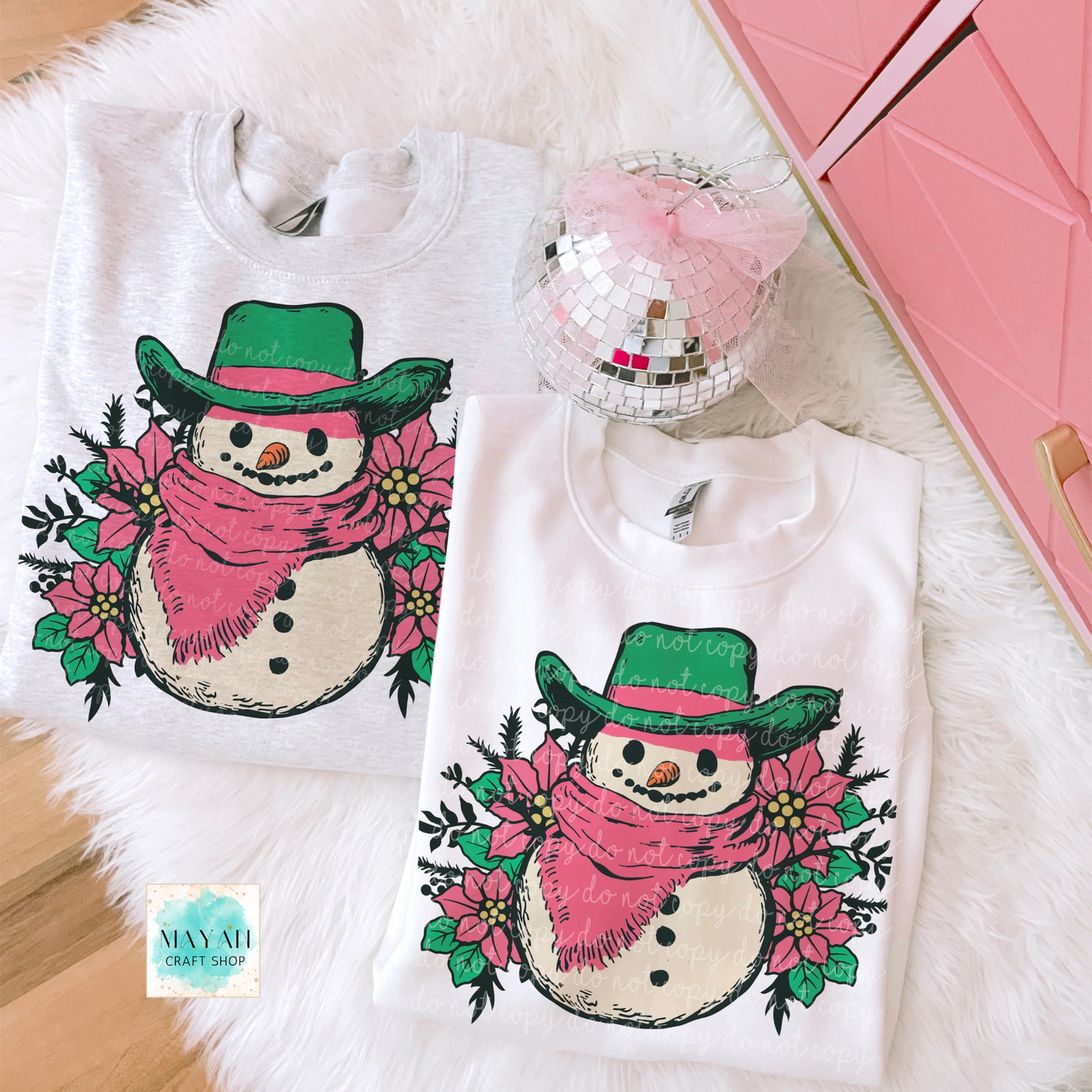 Two sweatshirts with a snowman design featuring a pink scarf and green hat on a fluffy white surface.