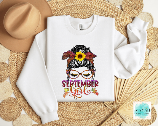 A white crewneck sweatshirt laid flat on a woven straw mat, decorated with a graphic of a woman's hair in a bun with a sunflower and fall leaves, and the text 'SEPTEMBER Girl' in colorful lettering. A tan hat and a folded paper fan are styled around it.