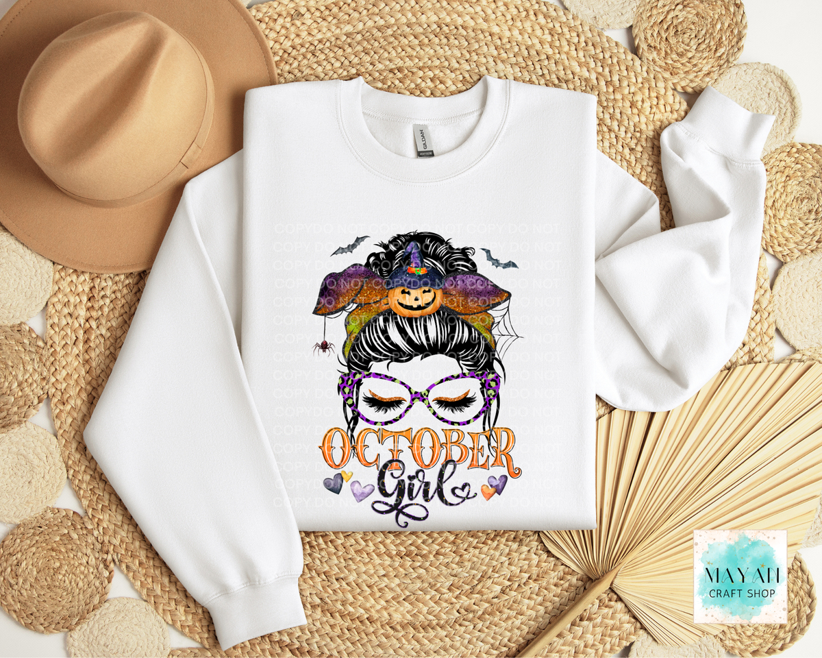 A white crewneck sweatshirt laid flat on a woven straw mat, featuring a colorful graphic of a witch's face with a pumpkin hat and the words 'OCTOBER Girl'. A tan hat.