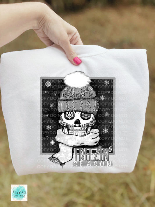 White sweatshirt with a skull wearing a winter hat and scarf design, held by a hand.