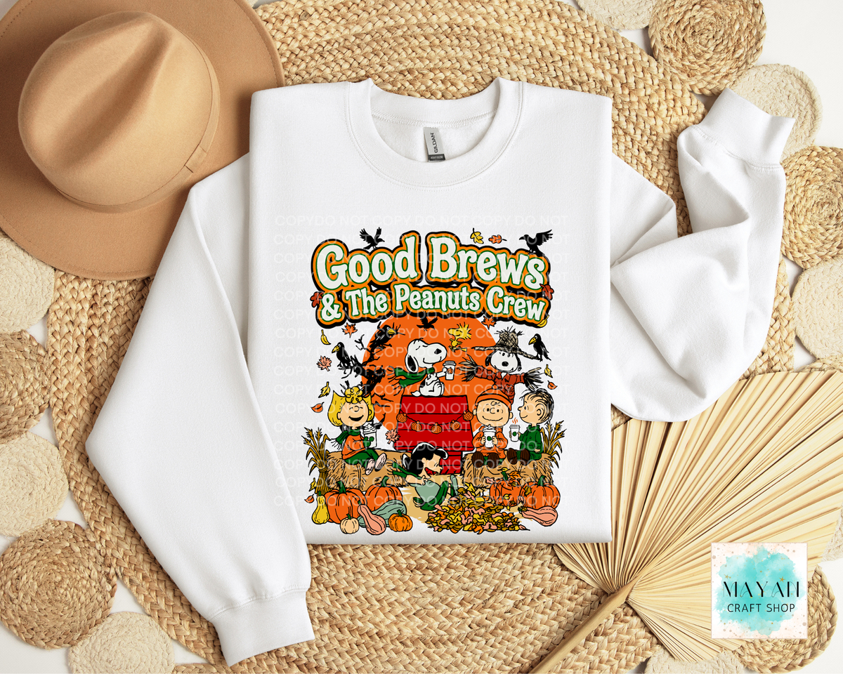 A white crewneck sweatshirt featuring a colorful 'Good Brews' autumn-themed graphic print, styled in a flat lay on a woven straw mat with a tan hat and dried palm leaves.