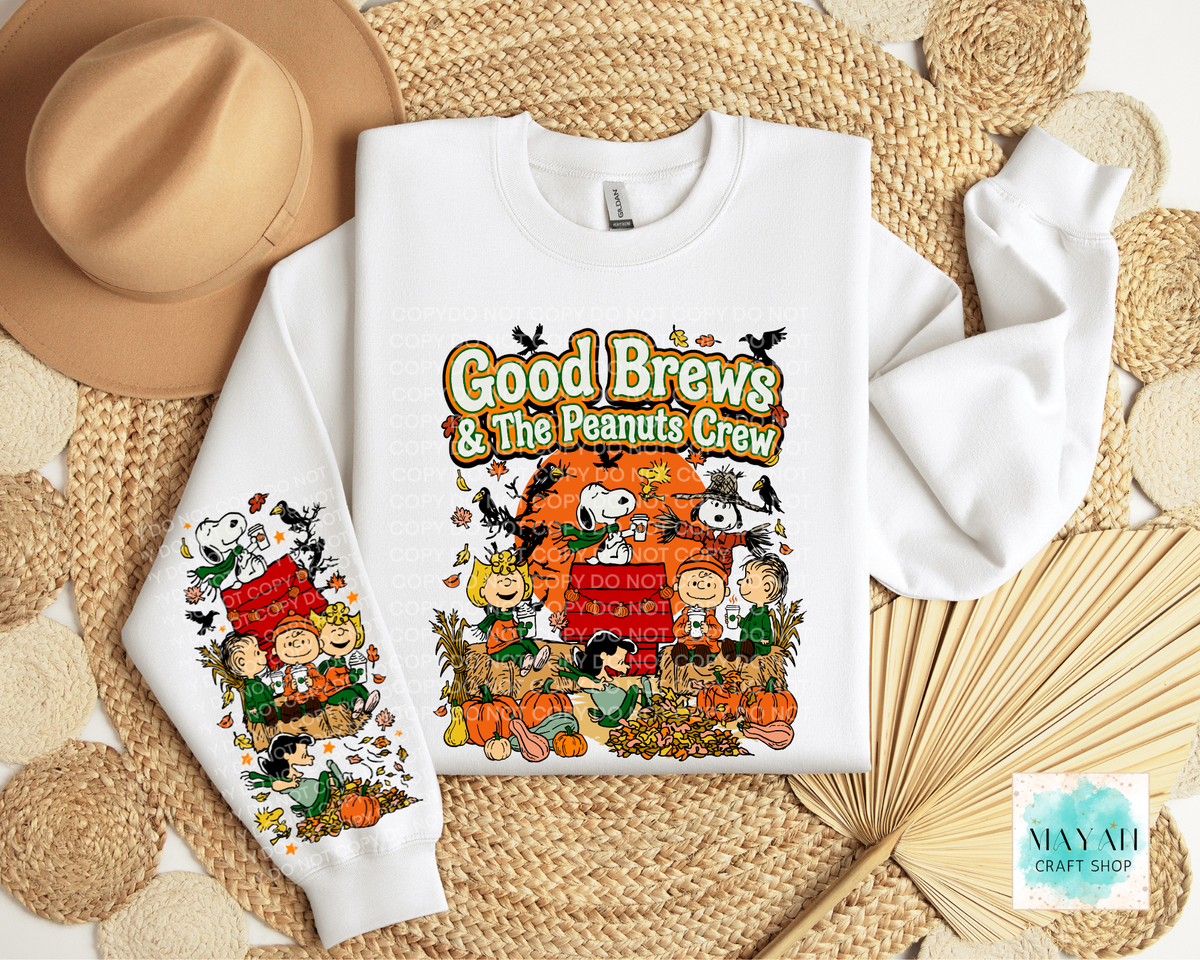 A folded white crewneck sweatshirt with a colorful 'Good Brews' graphic, styled in a flat lay with a tan hat, woven mat, and dried palm leaves.
