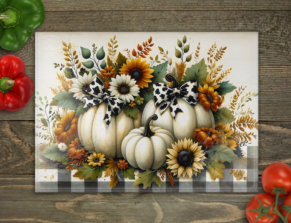 A cutting board featuring a fall harvest design with white pumpkins, sunflowers, leaves, and black-and-white bows, displayed on a rustic wood surface with red bell peppers and tomatoes around it.