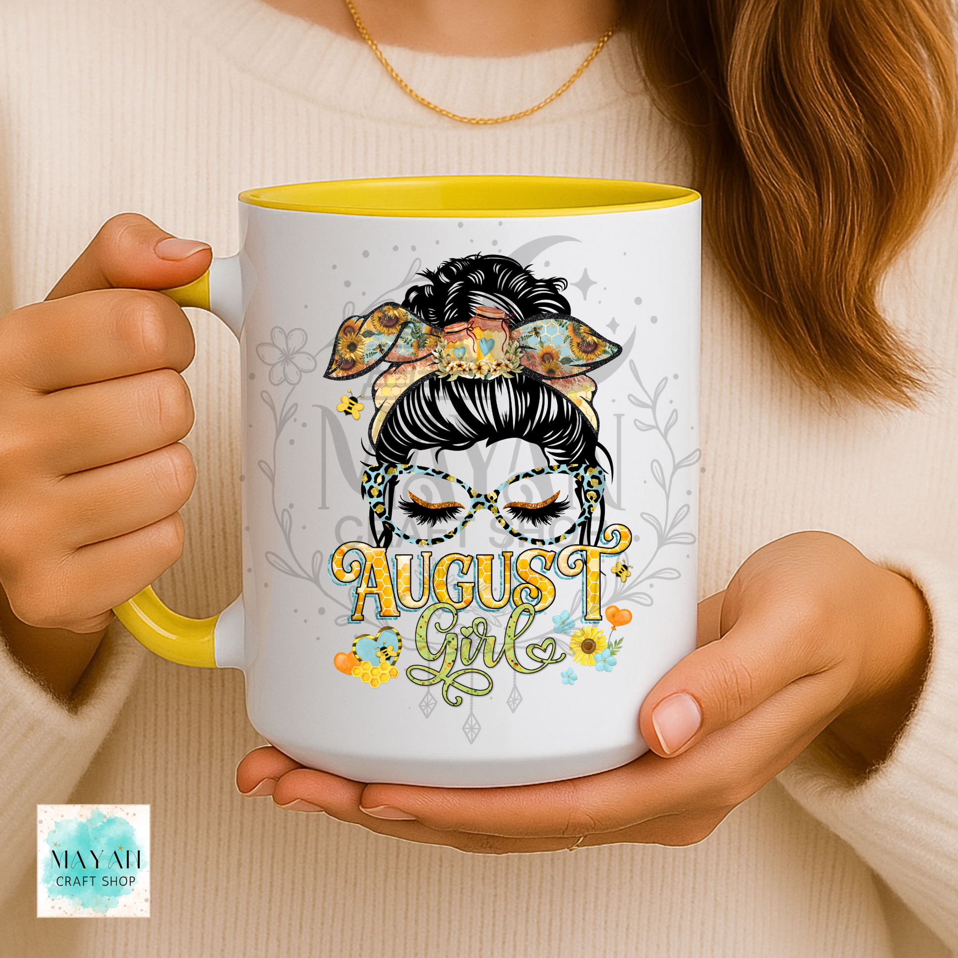 A person holds a white ceramic mug with a yellow interior and handle. The mug features a decorative illustration of a woman with sugar skull makeup, a floral headband, and the text 'August Girl' in yellow and green script.