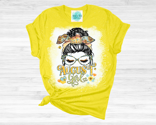 A bright yellow graphic t-shirt featuring a messy bun girl design and the text 'August girl' on a white wood background.