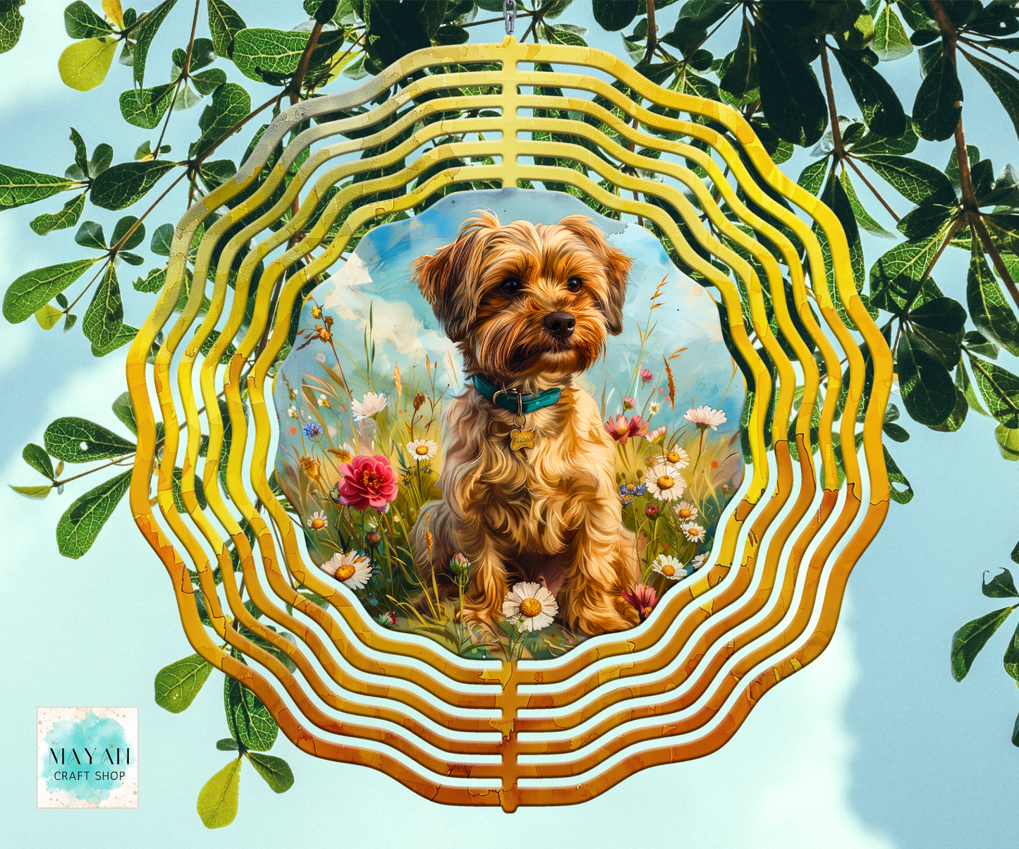 A wind spinner featuring a central painted portrait of a small brown dog sitting in a flower field. The piece is photographed hanging against a real outdoor background with green leaves and blue sky visible.