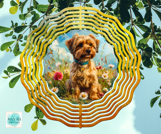 A wind spinner featuring a central painted portrait of a small brown dog sitting in a flower field. The piece is photographed hanging against a real outdoor background with green leaves and blue sky visible.