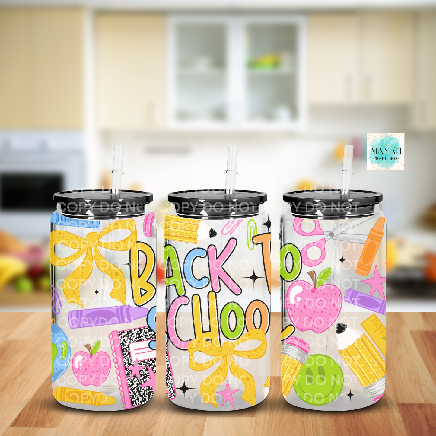 Three frosted glass cans with black lids and straws, decorated with colorful 'Back to School' graphics including yellow bows, pink apples, and multicolored stationery, displayed on a wooden table with a blurred kitchen background.